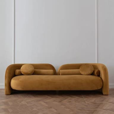 An asymmetrical daybed upholstered in mustard yellow velvet, featuring a distinct rounded, continuous back and armrest on one side that gradually lowers and disappears on the other. It includes two coordinating bolster pillows and one round cushion. — 3D model