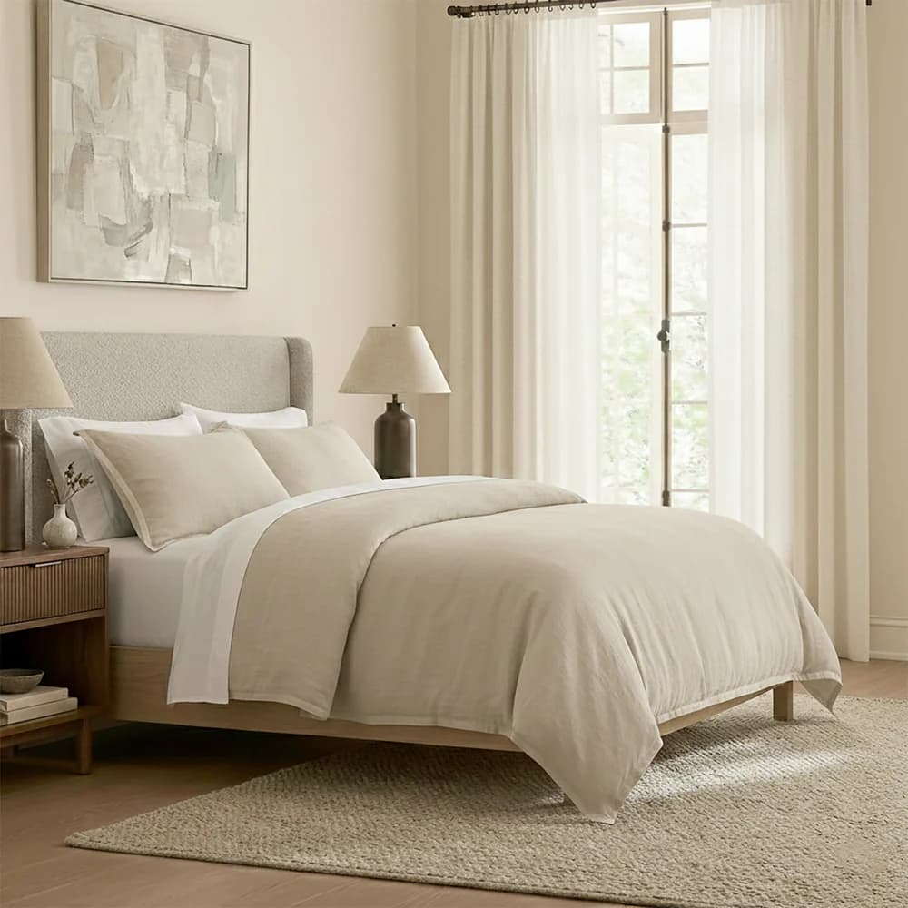 A bedroom scene featuring a bed, nightstand, artwork, and window treatments. The bed has a light-colored upholstered headboard and a duvet cover in a neutral tone. A wooden nightstand with ribbed drawer fronts is positioned to the left of the bed. An abstract painting hangs on the wall above the headboard. Sheer curtains cover a window behind the bed. — 3D model