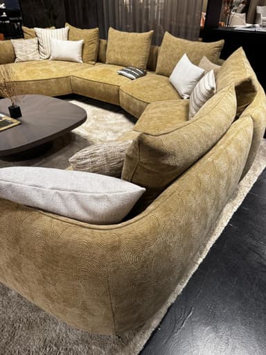 A sectional sofa with a textured, mustard-yellow fabric upholstery. The sofa features a low profile and deep seating. It is adorned with several throw pillows, including one in a neutral tone and others with subtle striped and textured patterns. The base of the sofa has a distinctive geometric pattern. Two dark wood coffee tables with rounded edges are positioned in front of the sofa, and a plush, light-colored rug covers the floor. — 3D model