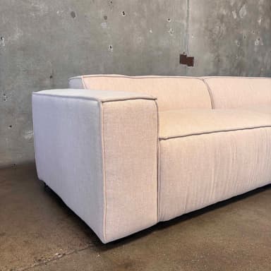 A modern, two-seater sofa with a low profile and blocky arms. The sofa is upholstered in a light-colored, textured fabric and features contrasting dark piping along the edges of the cushions and frame. The base appears to be a dark, solid plinth. It is positioned against a rough, concrete wall with visible imperfections and mounted fixtures. — 3D model