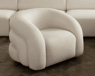 A contemporary armchair featuring a distinctive, fully upholstered design with rounded, volumetric forms. The chair is covered in a cream-colored, textured boucle fabric, giving it a soft and inviting appearance. It has integrated arms and a continuous base, appearing to rest directly on the floor without visible legs. — 3D model