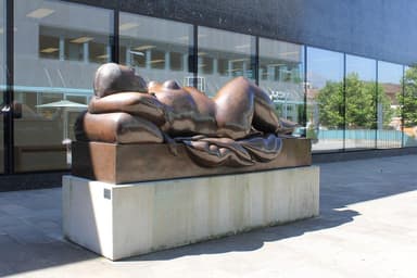 A large bronze sculpture depicting a reclining female figure, situated outdoors on a substantial concrete plinth in front of a modern glass building. The figure has soft, rounded forms and is posed with her head resting on a raised form resembling a pillow, with her body gently draped. — 3D model