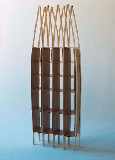 A tall, slender, modular shelving unit constructed primarily from light-colored plywood. It features multiple vertical sections, each with numerous small cubbies for storage or display. The top portion of the unit is characterized by a distinctive, open, slatted wooden framework, adding a unique architectural element. The entire unit is supported by slender, splayed wooden legs. — 3D model