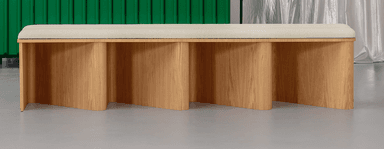 A long, low bench with a light wood finish. The bench features a flat top surface with slightly rounded edges. The base is composed of four vertically oriented, curved panels that create a wave-like or fluted effect. The overall design is minimalist and modern. — 3D model