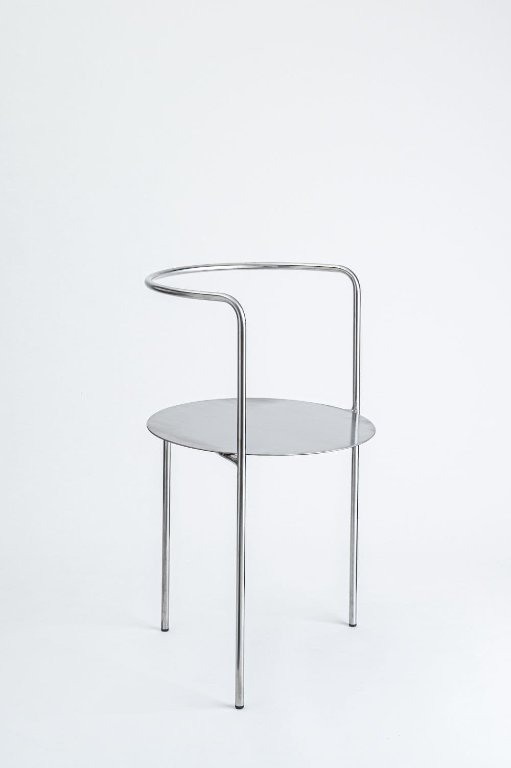 A minimalist chair constructed from bent metal tubing for the frame and legs, supporting a circular seat made of sheet metal. The frame consists of two vertical supports originating from the front legs and curving upwards and inwards to form a partial semi-circular backrest. The backrest is open at the top. The overall aesthetic is modern and sculptural. — 3D model