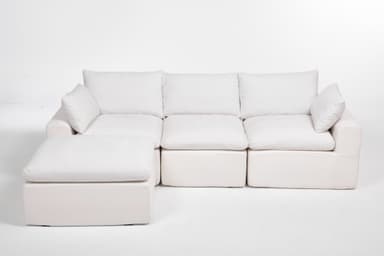 A modular sectional sofa with an attached ottoman. The sofa features deep, plush cushions and a low profile. It is upholstered in a textured white fabric. The configuration shown includes a corner section, two seat cushions, and an ottoman. The design is contemporary and emphasizes comfort. — 3D model
