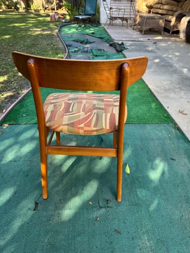 A close-up, overhead view of the padded seat and upper back support of an upholstered chair. The seat is covered in a multi-colored woven fabric featuring an abstract geometric pattern of wavy lines and rectangular blocks in shades of red, pink, beige, light green, and brown. Portions of the chair's wooden frame, including parts of the legs and back support, are visible, suggesting a medium-toned wood construction. The chair is positioned on a worn, teal-colored outdoor carpet or mat. — 3D model