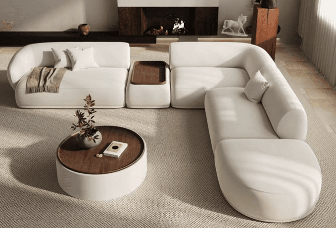 A multi-piece modular sectional sofa upholstered in a light, neutral fabric, featuring rounded edges and a low profile. One section includes a built-in wooden tray table. The arrangement is complemented by a separate, matching round coffee table with a wooden top. — 3D model