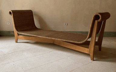A contemporary chaise lounge with a sinuous, ergonomic design. The frame is constructed from light-colored wood, featuring gently curved legs and supports. The seating surface and backrest are made of a woven material, possibly rattan or cane, creating a textured appearance. The headrest area is formed by a continuous, rolled curve of the woven material. — 3D model