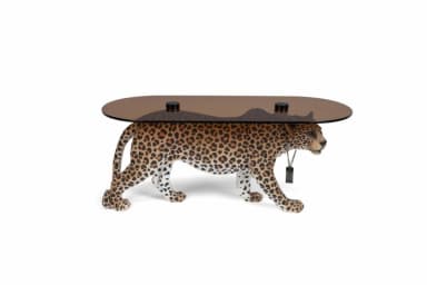 An oval coffee table supported by a realistically rendered leopard sculpture. The tabletop is made of smoked glass and is supported by black cylindrical posts that emerge from the leopard's back. The leopard sculpture is detailed with spotted fur patterns and appears to be in a walking pose. A black tag-like object is suspended from a chain around the leopard's neck. — 3D model