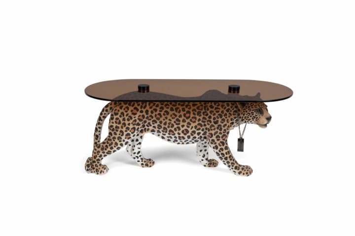 An oval coffee table supported by a realistically rendered leopard sculpture. The tabletop is made of smoked glass and is supported by black cylindrical posts that emerge from the leopard's back. The leopard sculpture is detailed with spotted fur patterns and appears to be in a walking pose. A black tag-like object is suspended from a chain around the leopard's neck. — 3D model