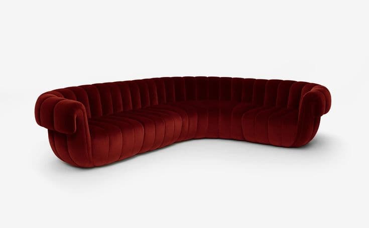 A large, elegantly curved sofa upholstered in deep red velvet, featuring prominent vertical channel tufting across its entire surface, including the back, seat, and rolled armrests. The sofa has a low-profile, monolithic appearance with no visible legs. — 3D model