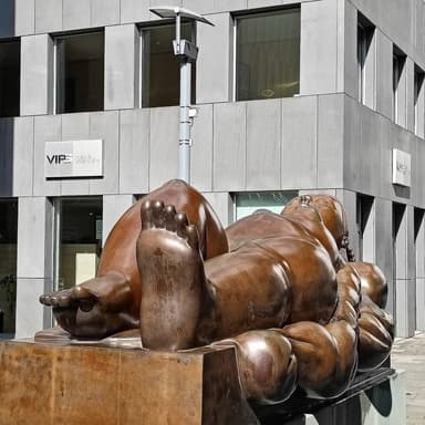 A large bronze sculpture depicting a reclining female figure, situated outdoors on a substantial concrete plinth in front of a modern glass building. The figure has soft, rounded forms and is posed with her head resting on a raised form resembling a pillow, with her body gently draped. — 3D model