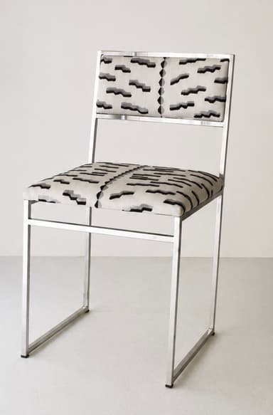 A contemporary armless chair featuring a minimalist, rectangular frame constructed from polished tubular metal. The seat and backrest are upholstered in a fabric with an abstract geometric pattern in shades of cream, black, and grey. The frame forms continuous loops for the legs and supports the seat and back. — 3D model