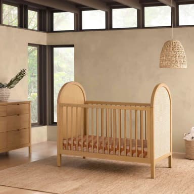 A wooden crib with a curved headboard and footboard. The sides feature vertical wooden slats, and one end panel is a woven cane material. A patterned mattress is visible inside. — 3D model