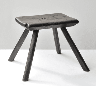 A rustic wooden stool with a square seat and three legs. The seat is flat with rounded edges and shows signs of wear, including some visible imperfections and grain texture. The legs are cylindrical and tapering slightly towards the bottom, appearing to be hand-shaped. The overall aesthetic suggests a primitive or country style. — 3D model