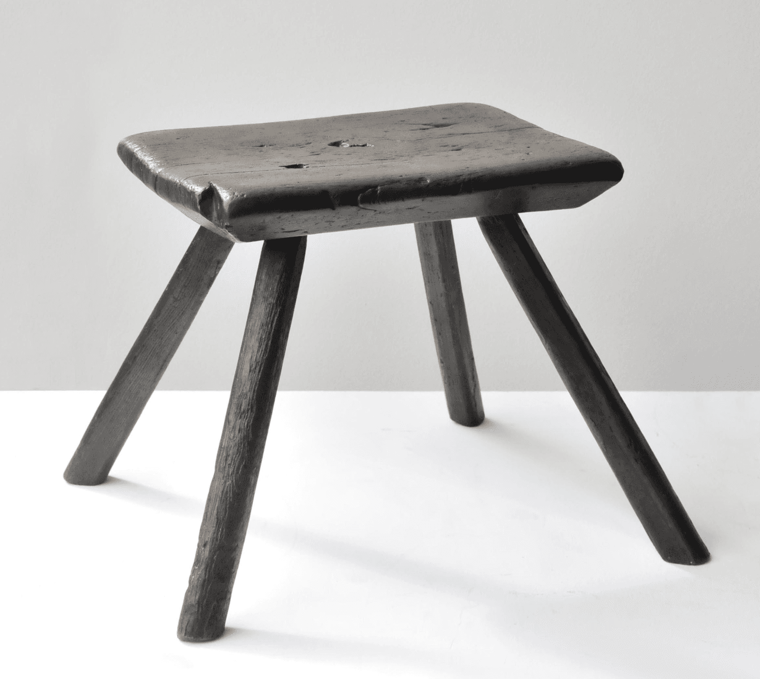 A rustic wooden stool with a square seat and three legs. The seat is flat with rounded edges and shows signs of wear, including some visible imperfections and grain texture. The legs are cylindrical and tapering slightly towards the bottom, appearing to be hand-shaped. The overall aesthetic suggests a primitive or country style. — 3D model