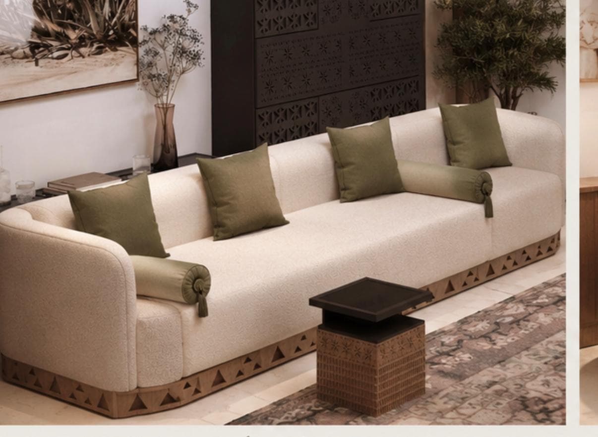 A contemporary three-seater sofa upholstered in light-colored bouclé fabric with rounded edges and a distinctive carved wooden base. It comes with four square and two bolster accent pillows in a contrasting olive green color. — 3D model