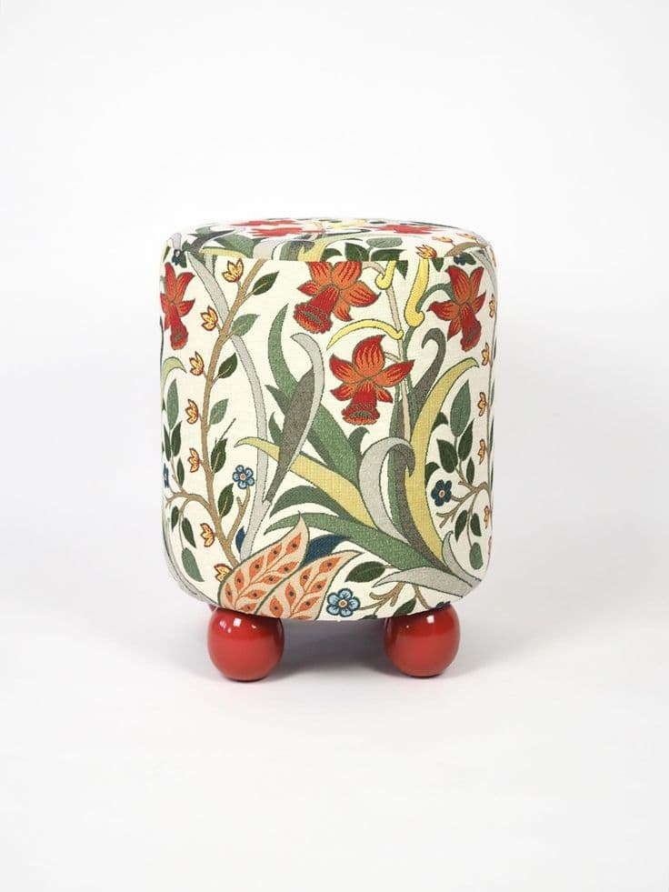 A compact, cylindrical ottoman upholstered in a multi-colored fabric featuring an intricate Arts and Crafts-inspired floral and botanical pattern. The main body of the ottoman rests on four short, spherical, brightly lacquered red feet, providing a contrasting base. The fabric design includes motifs of red flowers, green leaves, and delicate tendrils on an off-white background. — 3D model