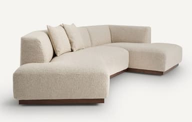 A modular sectional sofa with a low profile and clean lines. The sofa features a textured fabric upholstery and a dark wood base. It is configured with a chaise lounge on the left and a corner section on the right, accompanied by two square throw pillows. — 3D model