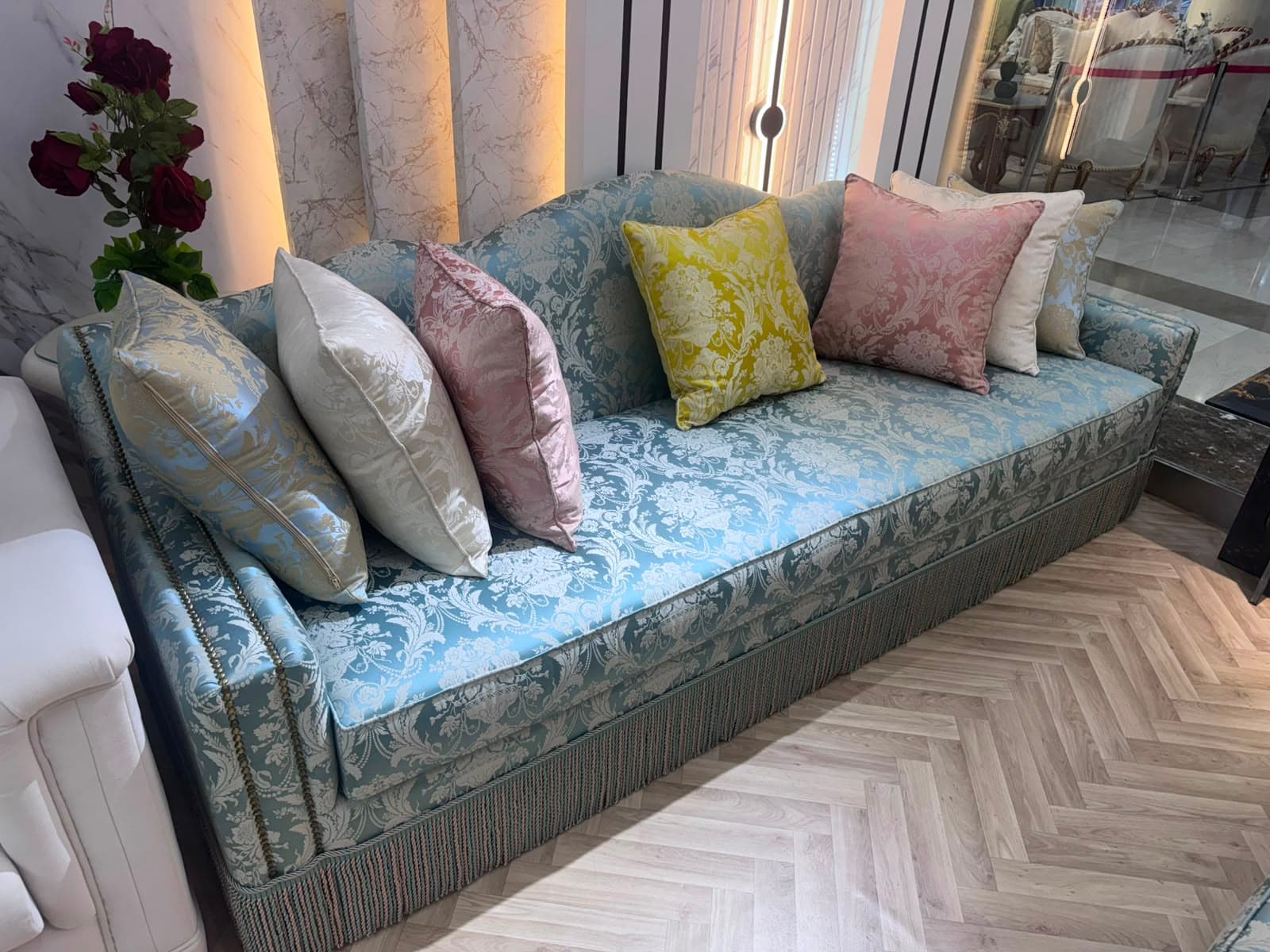 An elegant upholstered sofa featuring a light blue damask fabric with an ornate floral pattern. The sofa has a curved camelback design and rolled arms. A decorative fringe trim runs along the entire bottom edge, serving as a skirt. The seating area includes a single, long cushion, and the sofa is adorned with an assortment of throw pillows in complementary colors like pink, yellow, cream, and blue, all with various patterned fabrics. — 3D model
