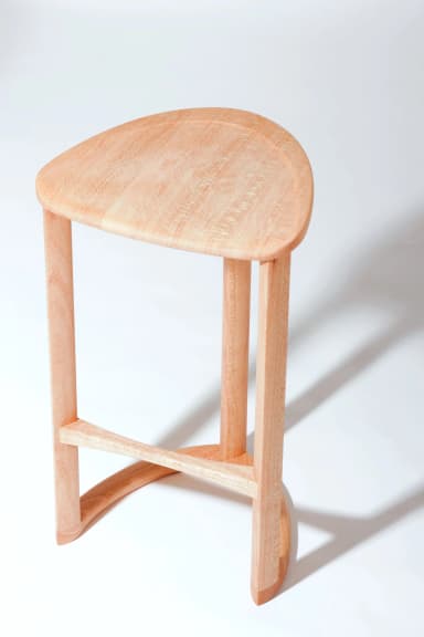 A wooden bar stool with a triangular, slightly dished seat and three cylindrical legs. One leg is positioned at the front and two at the back. A curved stretcher connects the two back legs near the bottom, and a diagonal stretcher connects the front leg to the rear left leg. The stool is constructed from light-colored wood with a visible grain. — 3D model