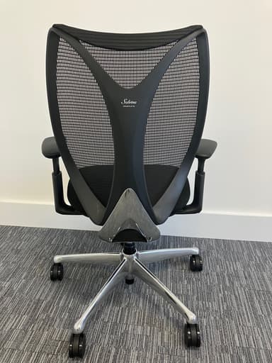 A modern ergonomic office chair is depicted from a frontal, slightly elevated perspective, set against a white wall and light grey carpeted floor. The chair features a black mesh backrest with a pronounced lumbar support curve, a black fabric-upholstered seat, and adjustable black plastic armrests. It is supported by a polished metallic five-star swivel base equipped with five black caster wheels. — 3D model