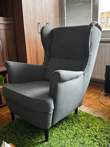 A contemporary wingback armchair upholstered in a textured grey fabric. The chair features a tall, slightly curved back with pronounced 'wings' at the top, a series of five decorative buttons forming a line down the center of the backrest, and sloped, rolled armrests. It has a removable seat cushion and stands on four slender, tapered dark-stained wooden legs. The overall design combines classic wingback elements with a modern, streamlined aesthetic. — 3D model