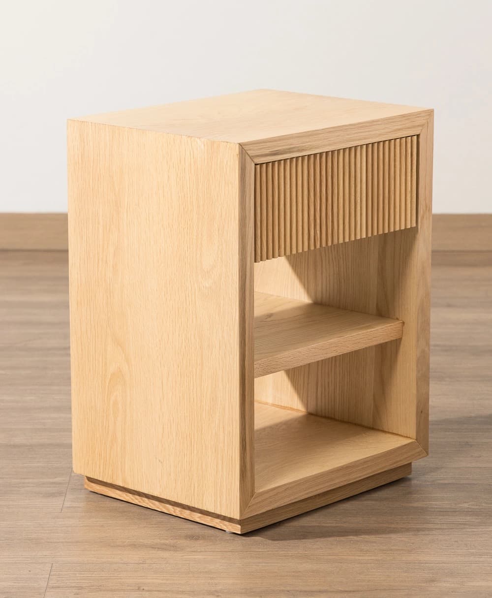 A modern nightstand crafted from light-toned wood, featuring a single top drawer with a distinct fluted or reeded facade, positioned above two open storage shelves. The unit rests on a recessed plinth base, giving it a slightly floating appearance. The overall design is minimalist and contemporary. — 3D model