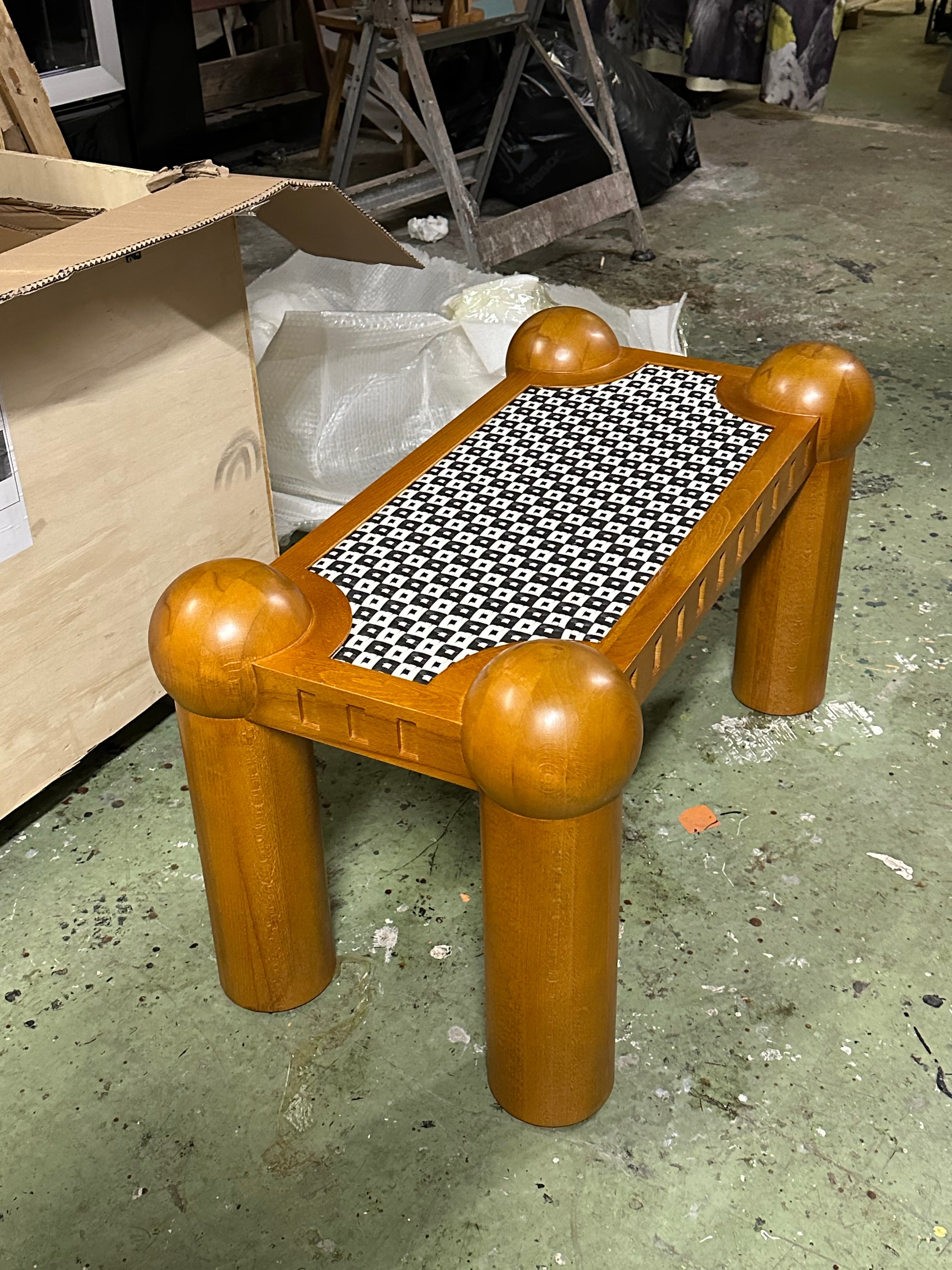 A postmodern-style accent table constructed from light brown wood, featuring a square top with a distinctive black and white patterned inlay. The table is supported by four chunky cylindrical legs, each topped with a large, smooth spherical finial at the corners of the tabletop. The apron beneath the tabletop exhibits small, rectangular carved details. — 3D model