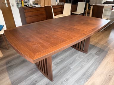 A modern dining table featuring a dark brown wooden top with elegantly curved edges and a matching slatted base. The table top displays a visible wood grain pattern, suggesting a veneer finish, and shows some minor surface wear. The base appears robust, designed with vertical wooden slats, which likely form a pedestal or trestle-style support. — 3D model