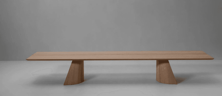 A long, rectangular dining table with a light wood finish. The tabletop appears to be constructed from multiple planks joined together, with a subtle seam running down the center. It is supported by two distinct, sculpted bases that taper from a wider curve at the bottom to a narrower point where they meet the underside of the tabletop. The bases have an angular, geometric yet softened aesthetic. The table is presented in a minimalist setting against a plain grey backdrop. — 3D model