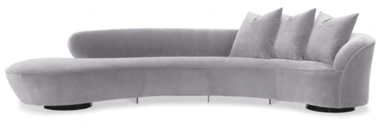 A contemporary, generously proportioned curved sofa upholstered in a light grey velvet fabric, featuring an integrated chaise lounge on the left side. The sofa exhibits a rounded back and armrest design, complemented by several loose back cushions. It is supported by low-profile black circular feet at its ends and slender, recessed metal supports along its central length. — 3D model