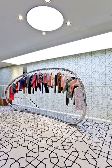 A modern, free-standing coat rack with a unique curved design. It features a polished metal frame that forms a semi-oval shape, supporting a horizontal rod along its upper curve. This rod is lined with multiple hangers displaying various garments. The base is a flat, elongated, curved metal piece that provides stability. The overall aesthetic is minimalist and sleek. — 3D model