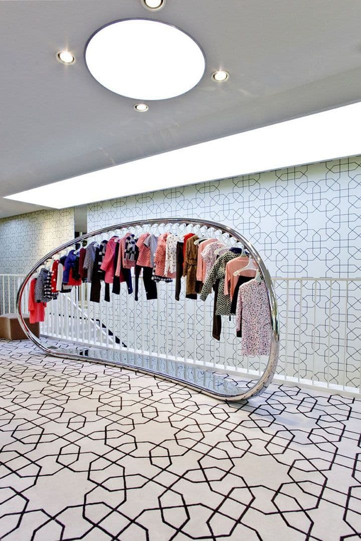 A modern, free-standing coat rack with a unique curved design. It features a polished metal frame that forms a semi-oval shape, supporting a horizontal rod along its upper curve. This rod is lined with multiple hangers displaying various garments. The base is a flat, elongated, curved metal piece that provides stability. The overall aesthetic is minimalist and sleek. — 3D model