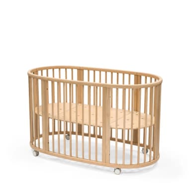 A natural wood, oval-shaped baby crib featuring vertical slats for safety and visibility, and small casters on the base for mobility. The mattress support is a slatted wooden panel. — 3D model
