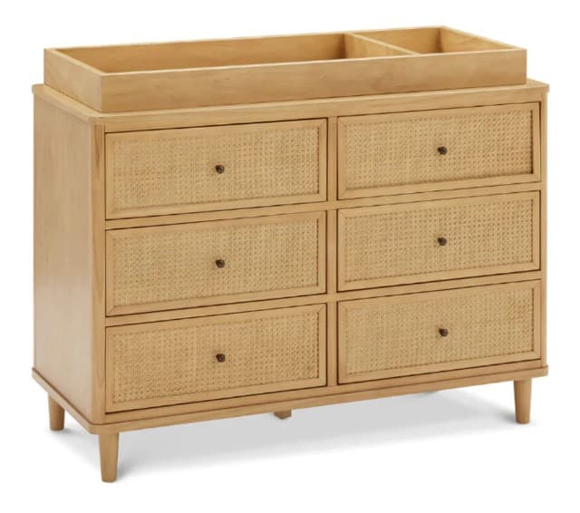A dresser with six drawers arranged in two columns of three. The drawer fronts feature a woven cane inset within a wooden frame. The dresser has a removable top unit with dividers, suggesting it may also function as a changing station. It is supported by four tapered legs. The overall construction appears to be wood with cane detailing. — 3D model