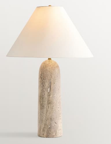 A contemporary table lamp featuring a solid, cylindrical base crafted from natural travertine stone, topped with a wide, conical fabric shade in a light cream or beige color. Small brass accents are visible where the shade connects to the base and at the top of the finial. — 3D model