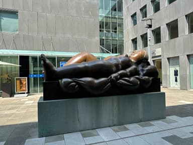 A large bronze sculpture depicting a reclining female figure, situated outdoors on a substantial concrete plinth in front of a modern glass building. The figure has soft, rounded forms and is posed with her head resting on a raised form resembling a pillow, with her body gently draped. — 3D model