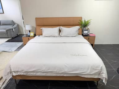 A modern queen-size bed with a tan upholstered headboard and frame, featuring two white pillows and white bedding. The bed is flanked by two matching brown nightstands with black glass tops, one of which holds a lamp, a decorative plant in a vase, and a book. The other nightstand is visible but less detailed. — 3D model