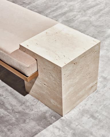 A contemporary bench featuring two rectangular, textured travertine stone bases supporting a horizontal upholstered seat. A thin, metallic frame, possibly brass or gold-toned, is visible beneath the cushion, connecting the two stone supports. The travertine exhibits natural pitting and veining characteristic of the material. The seat cushion is a neutral, light-colored fabric. — 3D model