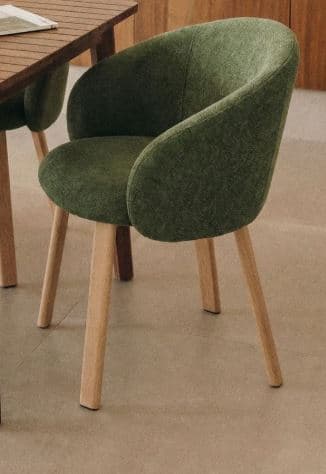 A contemporary dining armchair featuring a dark green, textured upholstered seat and back, with integrated armrests forming a continuous, rounded bucket shape. The chair is supported by four natural light-toned wooden legs that taper slightly downwards. The overall design suggests a comfortable yet minimalist aesthetic, suitable for modern interiors. — 3D model
