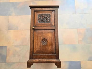 A small, dark brown wooden cabinet featuring a single door adorned with two distinct ornate carvings, equipped with a brass knob, and standing on a plinth base. — 3D model
