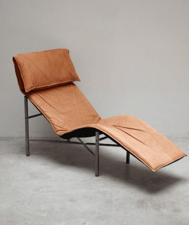 A modern chaise lounge with a thin, tubular metal frame supporting a flexible, reclining seat and back. The upholstery appears to be a tan-colored, possibly leather or suede, material with visible stitching and tufting in the seat area. A separate headrest cushion is positioned at the top of the chaise. The frame has a matte silver or grey finish. — 3D model