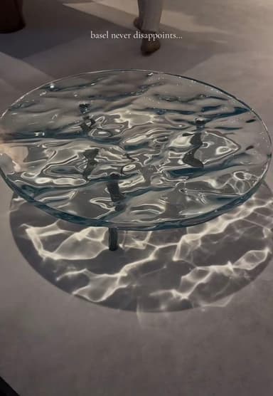 A round coffee table with a unique top designed to mimic the appearance of rippling water. The surface has an undulating texture, creating patterns of light and shadow on the floor beneath. It is supported by slender, dark-colored legs. — 3D model