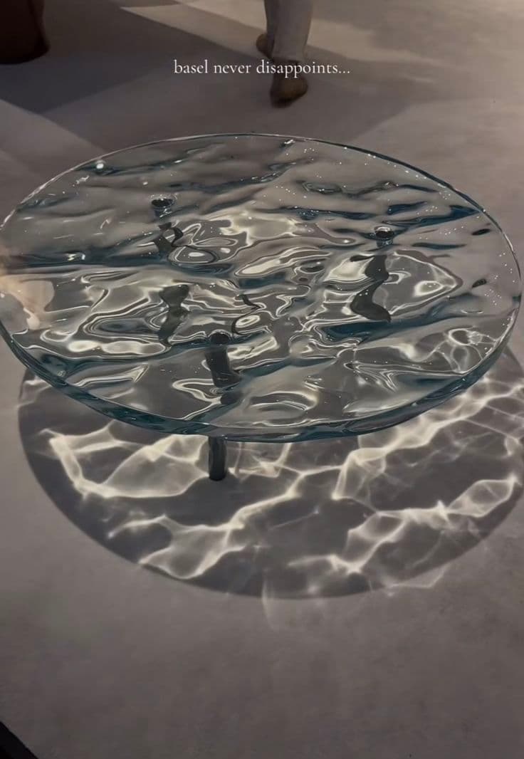 A round coffee table with a unique top designed to mimic the appearance of rippling water. The surface has an undulating texture, creating patterns of light and shadow on the floor beneath. It is supported by slender, dark-colored legs. — 3D model