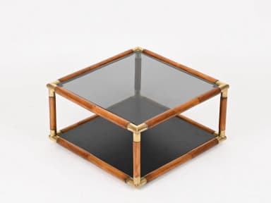 A square, two-tiered coffee table featuring a frame constructed from bamboo-style wood and joined by polished brass corner connectors. The top surface is a rectangular panel of smoked glass, while the lower shelf is a solid, opaque black surface, providing additional storage or display space. The design exudes a mid-century modern or Hollywood Regency aesthetic. — 3D model