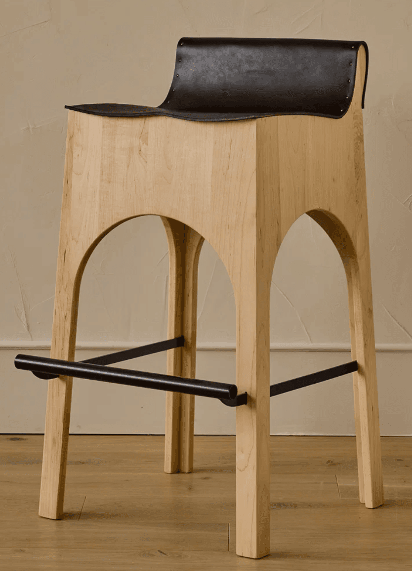A modern bar stool featuring a light-toned wooden frame with distinctive arched cutouts forming the legs. The seat and partial backrest are crafted from a single piece of dark, possibly black, leather, which is draped over the wooden frame and secured with visible rivets along the edges. A black metal footrest bar is integrated into the lower section of the stool's legs. — 3D model