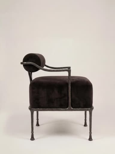 A robust armchair featuring a dark, possibly black or very dark brown, velvet upholstered seat and a cylindrical backrest. The frame is constructed from dark, textured, possibly forged iron or a similar heavy metal, exhibiting a somewhat raw and organic aesthetic. The legs are slender but robust, tapering slightly to small, disc-like feet. The backrest is suspended above the seat, supported by the frame's uprights, with a small V-shaped support element visible at the rear. — 3D model