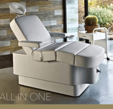 A light gray adjustable hydraulic treatment chair, designed for spa, salon, or medical aesthetic applications. It features segmented upholstered cushions for the backrest, seat, and leg rest, allowing for multi-position recline. The headrest is adjustable and includes a removable face cradle. Padded armrests are present, with one featuring an attached cup holder. The chair is supported by a robust, solid, light gray base, likely housing hydraulic or electric adjustment mechanisms. — 3D model